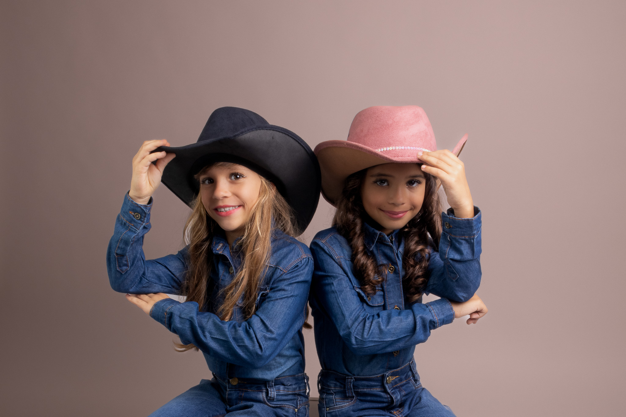 Todos pela acessibilidade: Manuella e Antonella estão sentadas, cada uma segurando delicadamente seu chapéu. A luz suave do estúdio destaca os detalhes de suas expressões calmas e confiantes. A foto captura um momento de descontração, onde a conexão entre as irmãs gêmeas é reforçada pela postura serena e o gesto sutil de segurar os chapéus, criando uma cena leve e elegante.