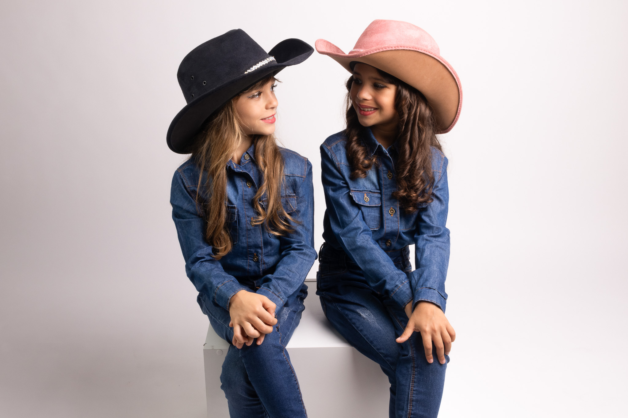 Todos pela acessibilidade: Manuella e Antonella trocam olhares sorridentes, refletindo a conexão única entre irmãs gêmeas. A luz suave do estúdio realça a expressão de afeto entre elas.