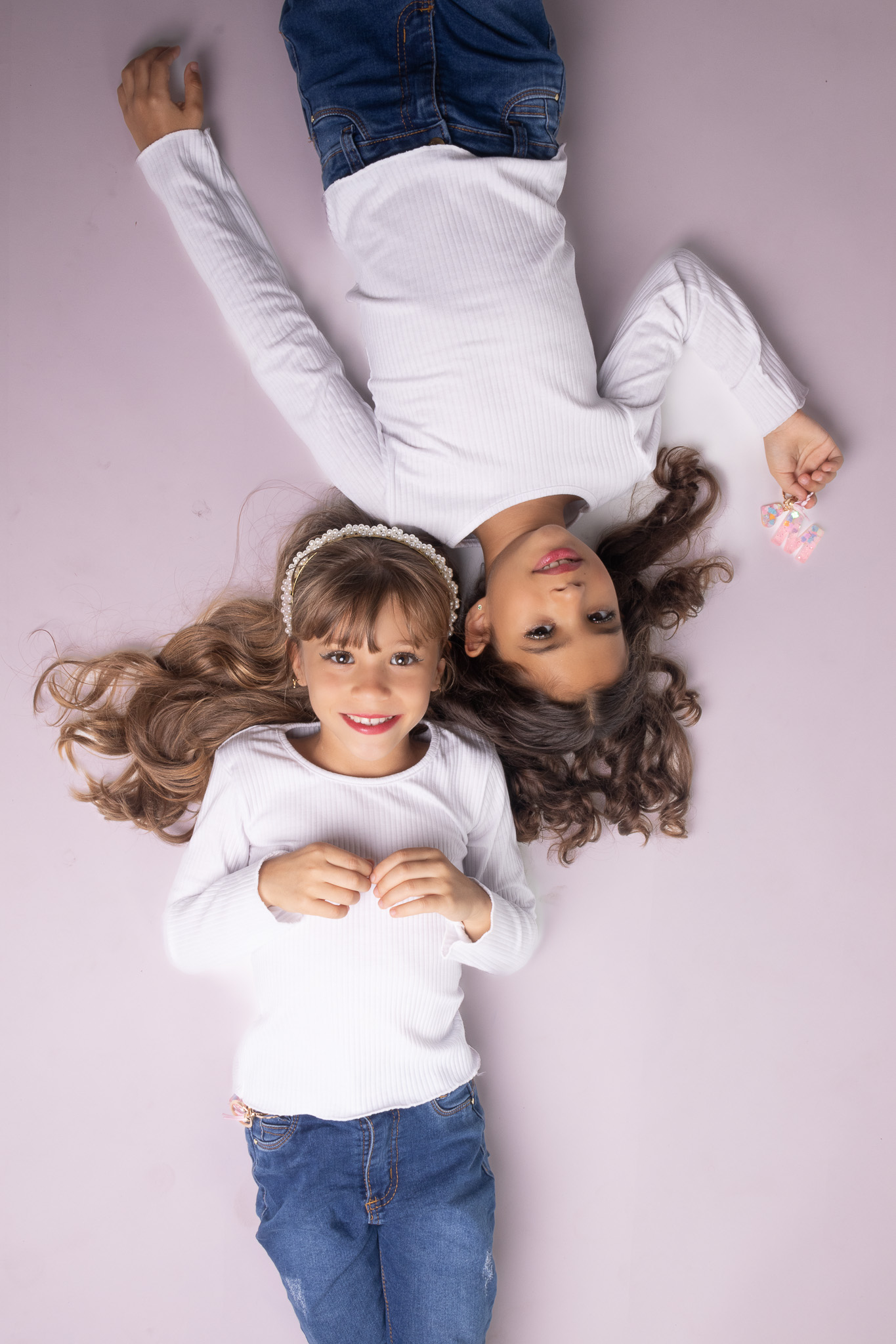 Todos pela acessibilidade: Manuella e Antonella estão deitadas lado a lado, capturadas de cima. A luz suave do estúdio cria uma atmosfera calma e aconchegante, destacando a simetria e conexão entre as irmãs gêmeas. Seus sorrisos leves refletem a serenidade e cumplicidade que compartilham nesse momento de descanso.