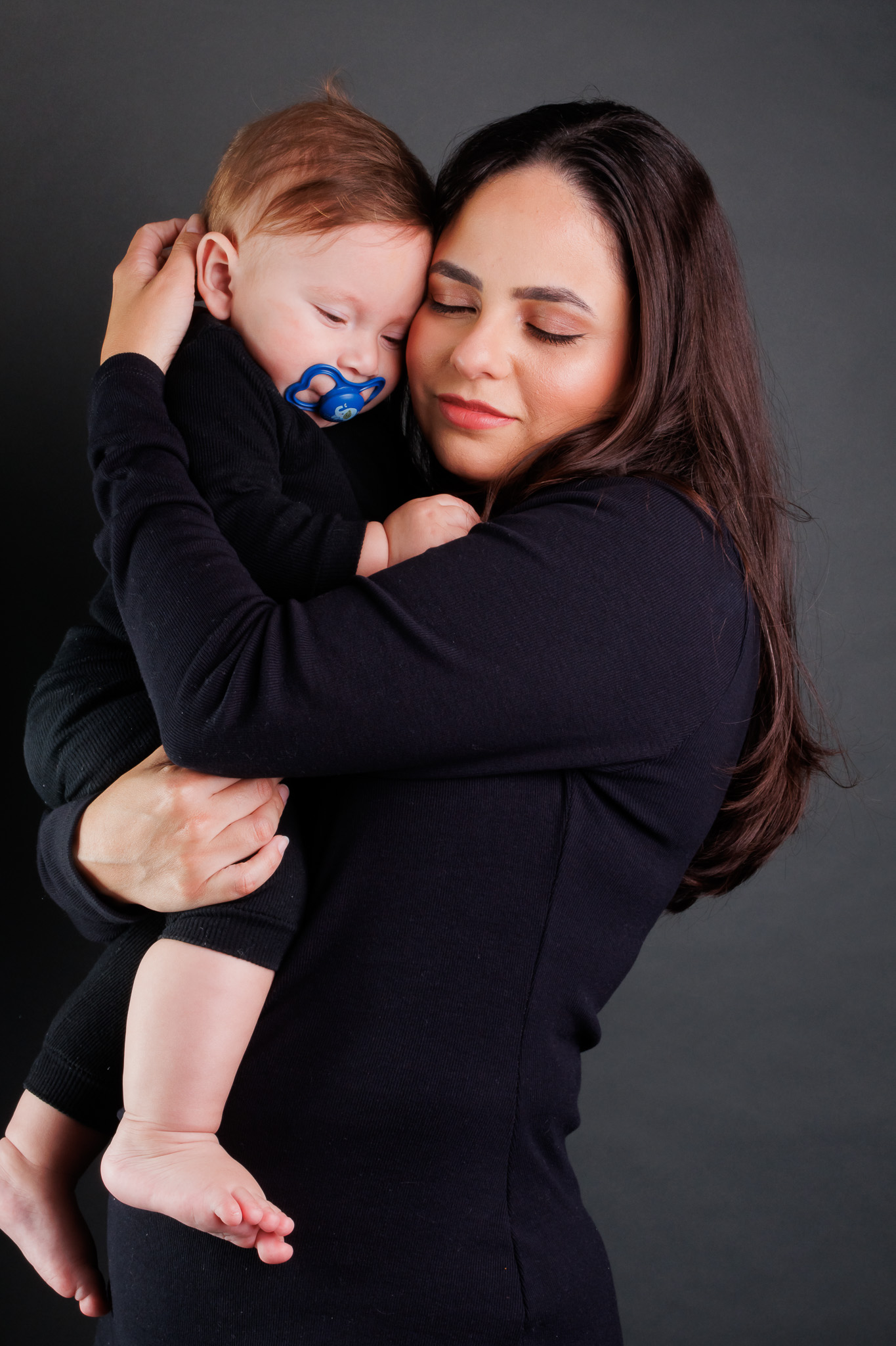 Todos pela acessibilidade: Mãe e filha estão em um abraço caloroso, ambas sorrindo, com a luz controlada realçando cada detalhe de suas expressões. A fotógrafa utilizou sua habilidade para criar uma atmosfera acolhedora no estúdio, destacando o carinho entre as duas com uma iluminação suave e bem equilibrada.